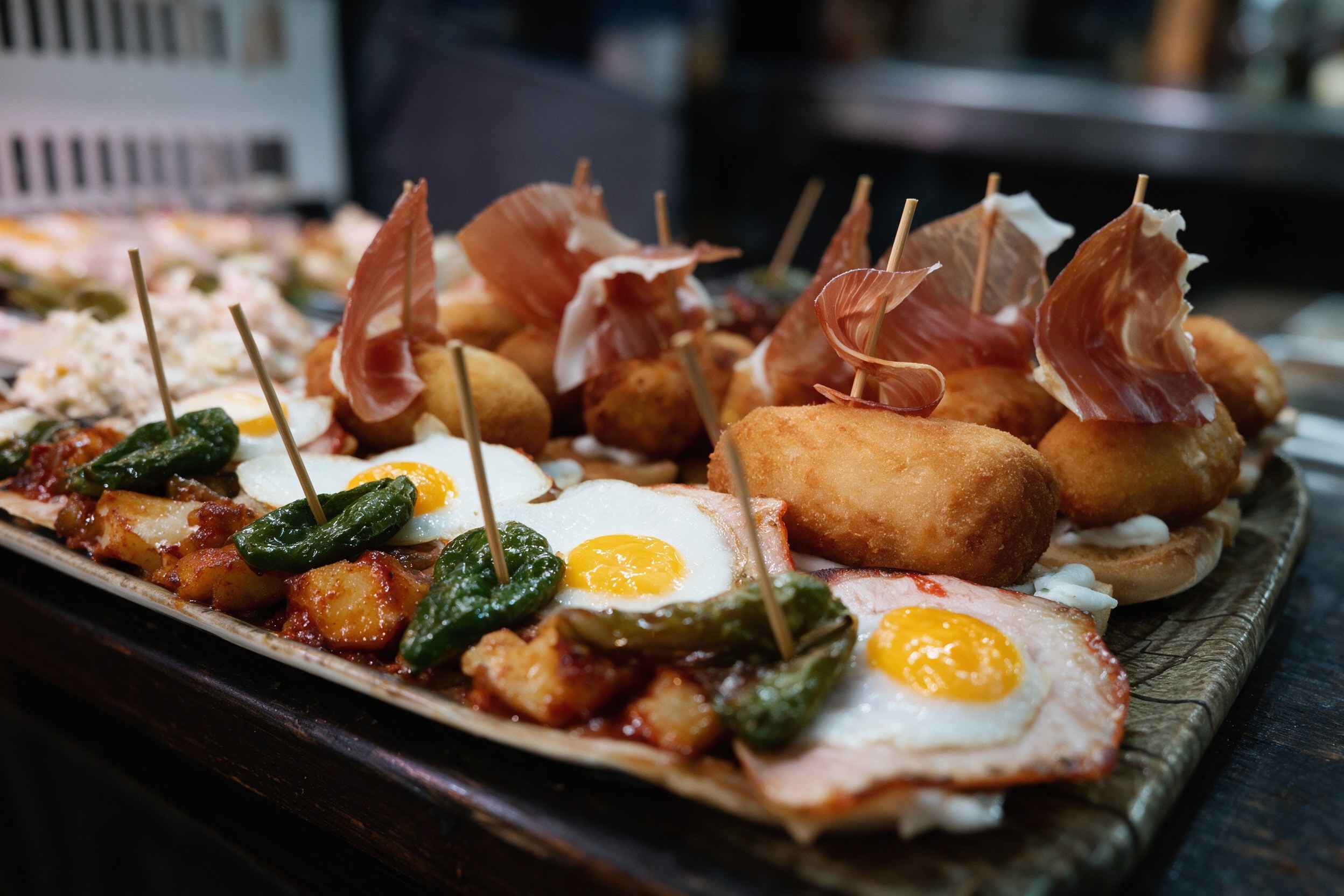 Bandeja larga de tapas variadas con croquetas, huevo frito, jamón, patatas y pimientos en la barra